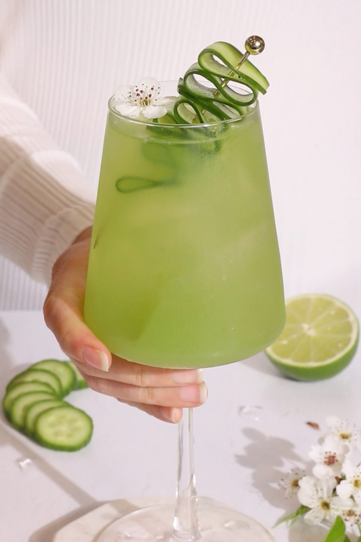 green mocktail with cucumber lime and elderflower in a wine glass garnished with a cucumber ribbon and a white flower.