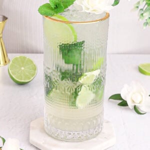 green and white non alcoholic elderflower cordial in a tall glass with a gold rim garnished with mint and white flower.