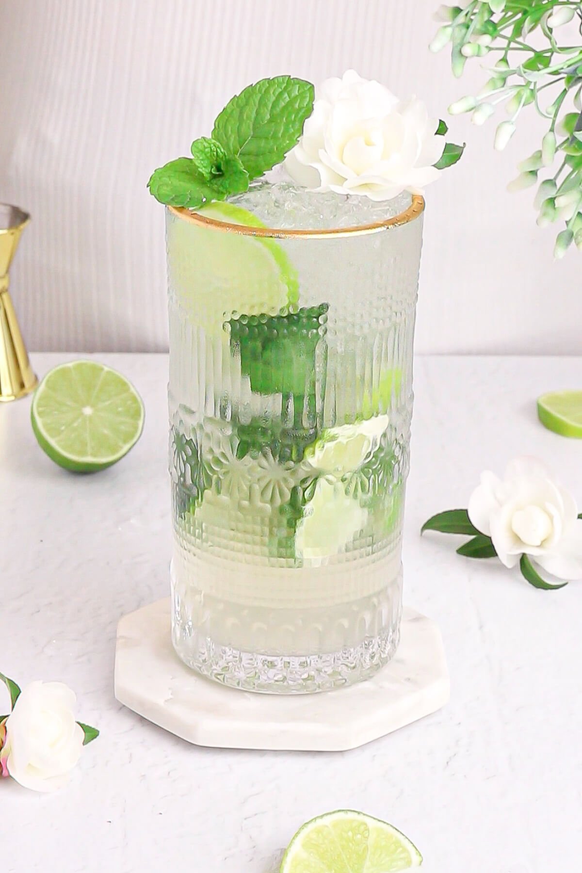 green and white non alcoholic elderflower cordial in a tall glass with a gold rim garnished with mint and white flower.