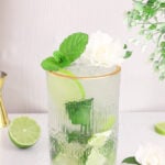 green and white elderflower mojito mocktail in a tall gold rimmed gladd with fresh lime and mint in the background.