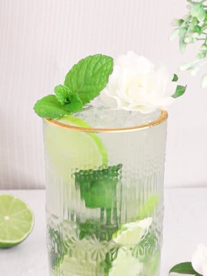 green and white elderflower mojito mocktail in a tall gold rimmed gladd with fresh lime and mint in the background.