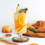 a non alcoholic apple cider drink in a tall glass with a gold rim garnished with rosemary pear apple and orange.