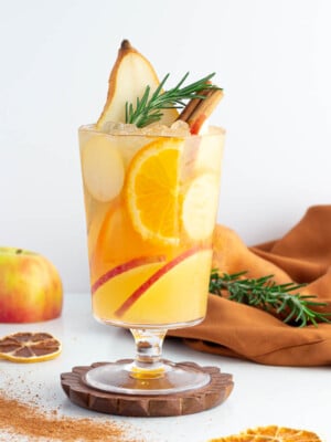 a non alcoholic apple cider drink in a tall glass with a gold rim garnished with rosemary pear apple and orange.