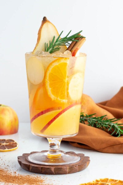 a non alcoholic apple cider drink in a tall glass with a gold rim garnished with rosemary pear apple and orange.