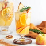 alcohol-free apple cider sangria in a tall glass garnished with orange, rosemary pear and cinnamon with a pitch of the sangria in the background.