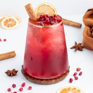 cinnamon mocktail in a short glass with a gold rim garnished with cinnamon pomegranate and orange