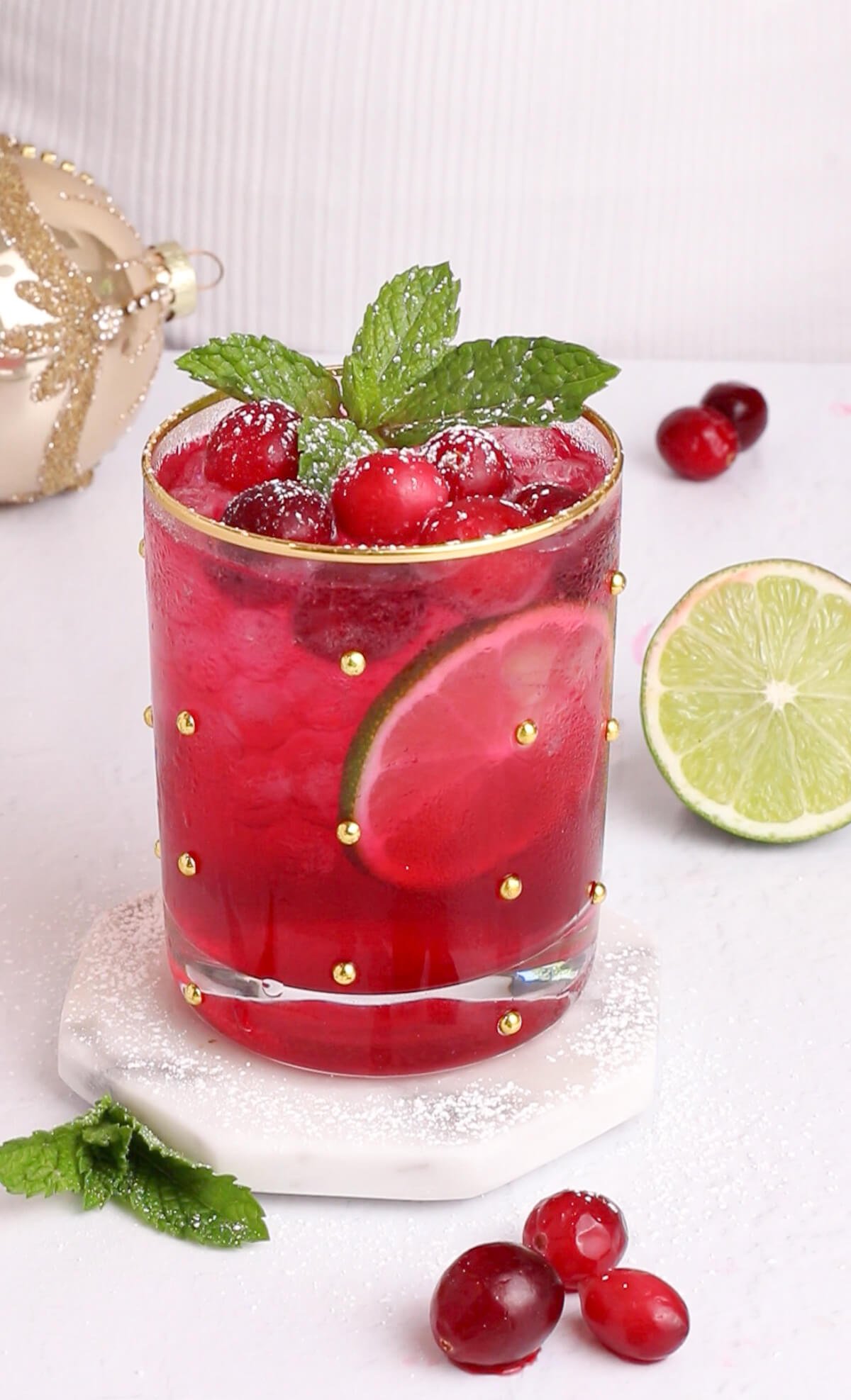 red cranberry holiday mocktail garnished with frozen cranberries mint and lime in a glass with a gold rim.