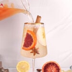 square image of a non alcoholic white sangria being poured into a wine glass with a gold rim.