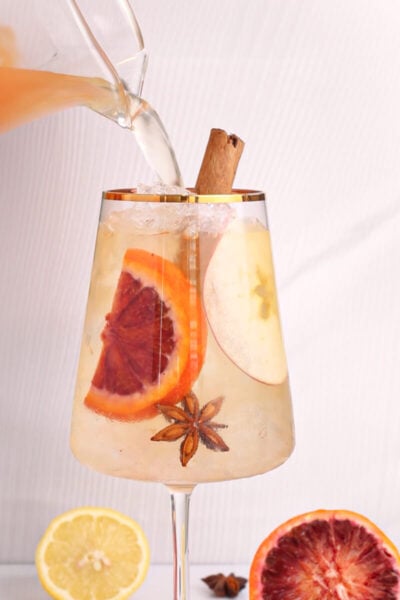 square image of a non alcoholic white sangria being poured into a wine glass with a gold rim.