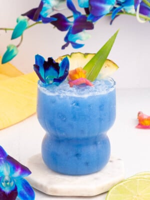 A vibrant blue Hawaii mocktail garnished with a slice of pineapple, an aloe leaf, and colorful flowers. It sits on a marble coaster, surrounded by lime slices and blue flowers, with a yellow cloth in the background.