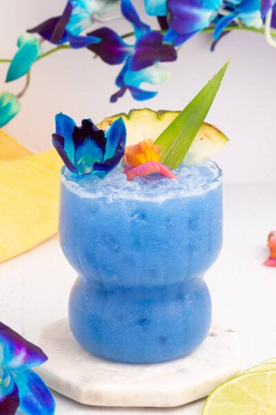 A vibrant blue Hawaii mocktail garnished with a slice of pineapple, an aloe leaf, and colorful flowers. It sits on a marble coaster, surrounded by lime slices and blue flowers, with a yellow cloth in the background.