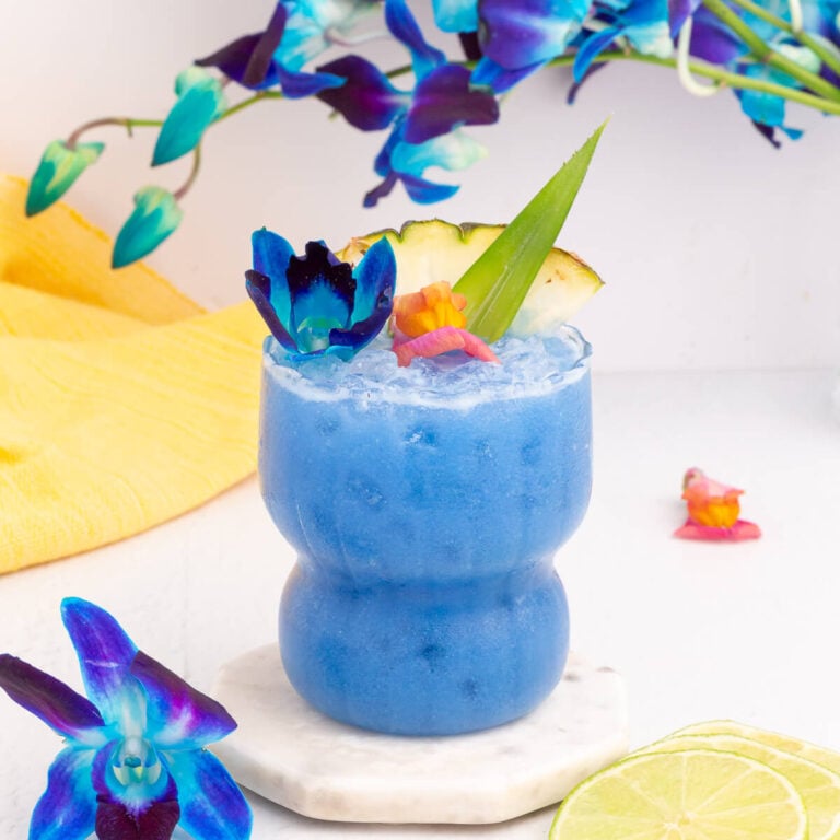 A vibrant blue Hawaii mocktail garnished with a slice of pineapple, an aloe leaf, and colorful flowers. It sits on a marble coaster, surrounded by lime slices and blue flowers, with a yellow cloth in the background.