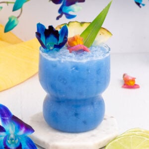 A vibrant Blue Hawaii mocktail in a curvy glass, garnished with colorful flowers, a pineapple slice, and a green leaf, sits on a marble coaster. Blue flowers and lime slices are nearby, with a yellow towel in the background.