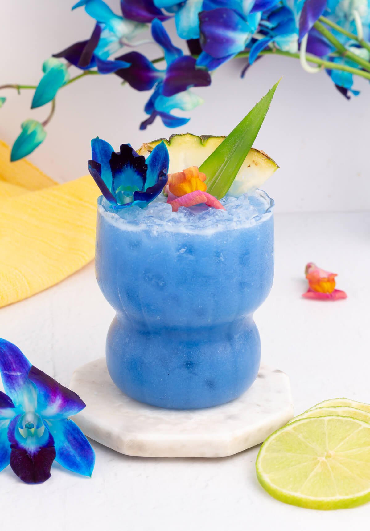 A vibrant Blue Hawaii mocktail in a curvy glass, garnished with colorful flowers, a pineapple slice, and a green leaf, sits on a marble coaster. Blue flowers and lime slices are nearby, with a yellow towel in the background.