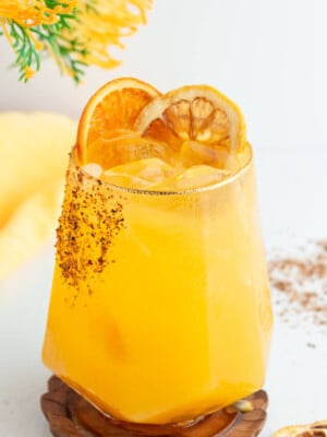 A refreshing yellow cocktail served in a geometric glass with ice, garnished with two dried orange slices. The rim is lightly coated with chili powder. A yellow cloth and a sprig of yellow flowers are in the background.