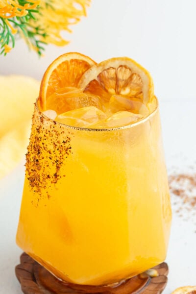 A refreshing yellow cocktail served in a geometric glass with ice, garnished with two dried orange slices. The rim is lightly coated with chili powder. A yellow cloth and a sprig of yellow flowers are in the background.