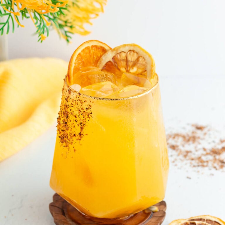 A refreshing yellow cocktail served in a geometric glass with ice, garnished with two dried orange slices. The rim is lightly coated with chili powder. A yellow cloth and a sprig of yellow flowers are in the background.