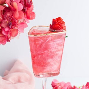 A pink, shimmery mocktail in a square glass with a gold rim, garnished with a strawberry. Pink flowers are in the background, along with a soft pink cloth.