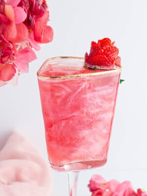A pink, shimmery mocktail in a square glass with a gold rim, garnished with a strawberry. Pink flowers are in the background, along with a soft pink cloth.