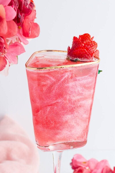 A pink, shimmery mocktail in a square glass with a gold rim, garnished with a strawberry. Pink flowers are in the background, along with a soft pink cloth.