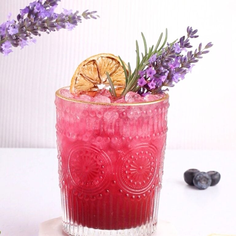 A decorative glass filled with a vibrant purple cocktail, topped with crushed ice. It is garnished with a sprig of lavender, rosemary, and a dried lemon slice. Two sprigs of lavender lean against the glass. Blueberries are nearby.