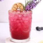 A vibrant pink drink in a textured glass sits on a white coaster. Its garnished with a dried lemon slice and sprigs of lavender. Lemon slices and blueberries are nearby, enhancing the fresh, floral presentation.