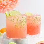 Two pink paloma mocktails with lime slices sit on marble coasters, surrounded by ice cubes. The glasses have rims coated with lime zest. In the background, a peach-colored flower and sliced grapefruit and lime add a vibrant touch.