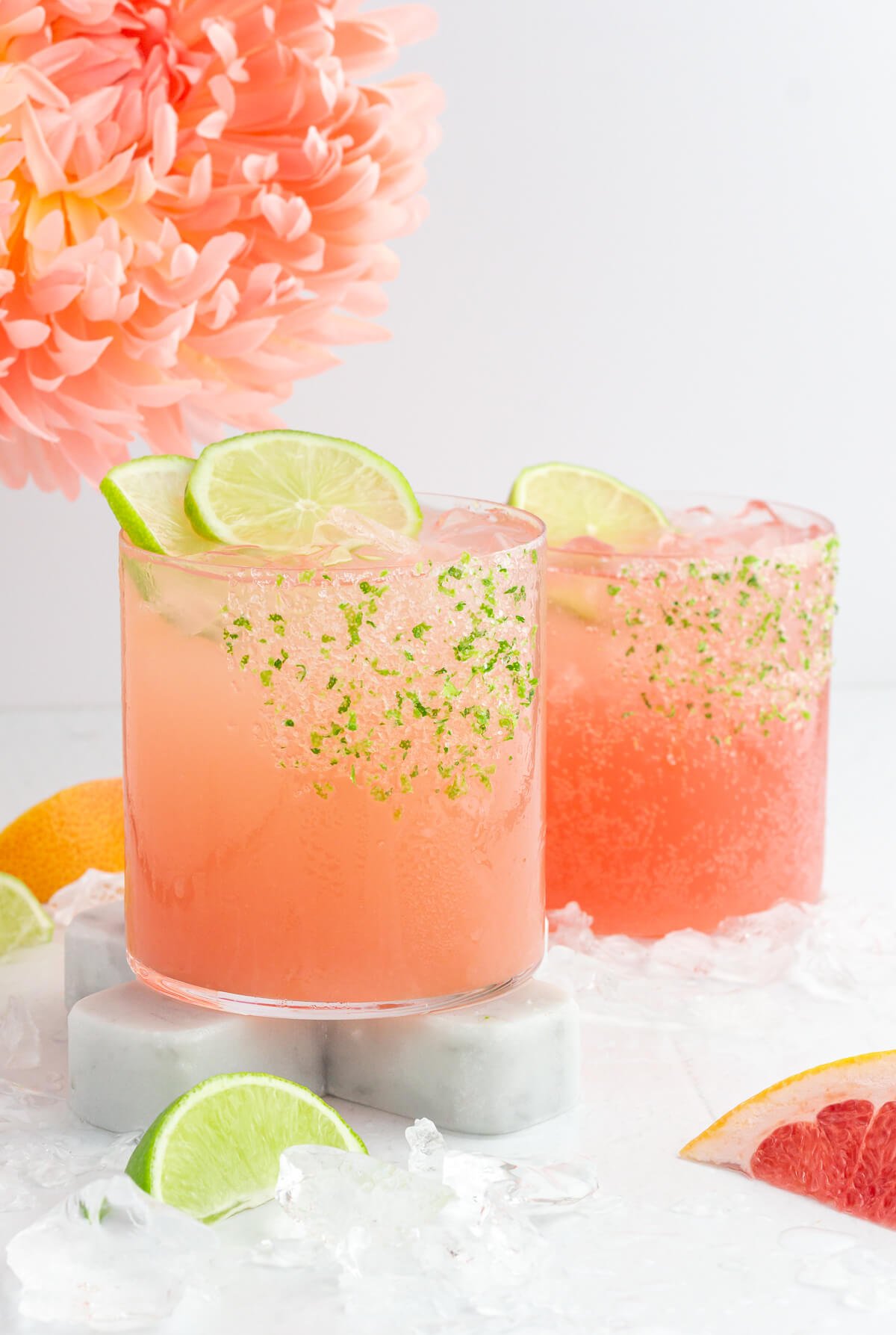 Two pink paloma mocktails with lime slices sit on marble coasters, surrounded by ice cubes. The glasses have rims coated with lime zest. In the background, a peach-colored flower and sliced grapefruit and lime add a vibrant touch.