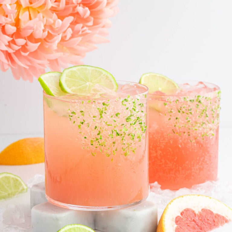Two non-alcoholic palomas with lime slices on glasses rimmed with mint, placed on stone coasters. Surrounding garnishes include lime wedges and grapefruit slices. A large peach flower is in the background. The scene is bright and refreshing.
