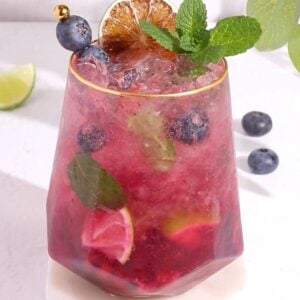 A glass of blueberry mojito moccktail garnished with fresh blueberries, a mint sprig, a slice of dried lime, and a gold-tipped pick. Ice, lime wedges, and mint are visible inside the pink drink. Blueberries and lime are scattered nearby.