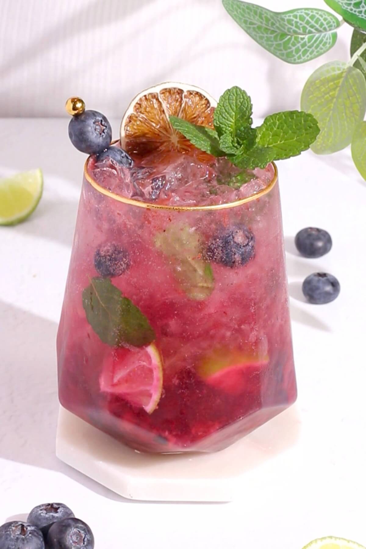 A glass of blueberry mojito moccktail garnished with fresh blueberries, a mint sprig, a slice of dried lime, and a gold-tipped pick. Ice, lime wedges, and mint are visible inside the pink drink. Blueberries and lime are scattered nearby.