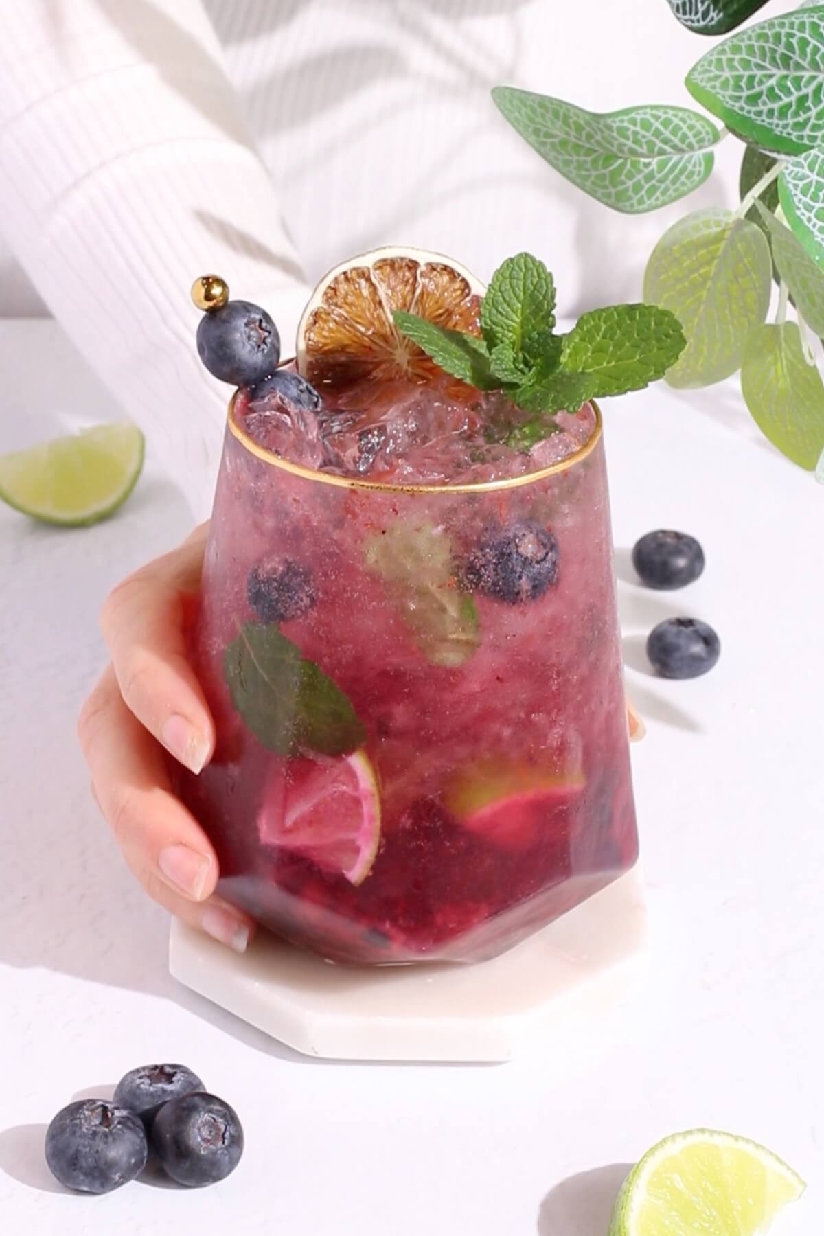 A hand holds a glass of a purple blueberry mocktail with ice, mint leaves, lime wedges, a dried lime garnish, and fresh blueberries, on a white surface with scattered blueberries and lime slices nearby.