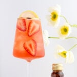 A glass of pink iced glowy skin spritz mocktail with sliced strawberries and a lemon wheel, set against a white background with white flowers and a small brown bottle nearby.
