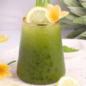 A glass of iced green matcha mocktail garnished with a lemon slice, yellow flower, and pineapple leaves, sits on a white surface with extra lemon slices and a pineapple in the background.