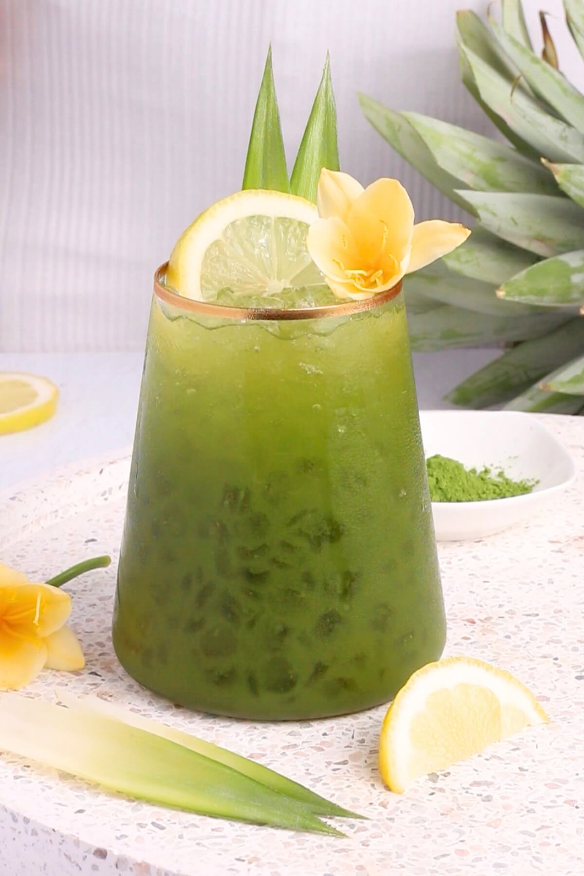 A glass of iced green matcha mocktail garnished with a lemon slice, yellow flower, and pineapple leaves, sits on a white surface with extra lemon slices and a pineapple in the background.