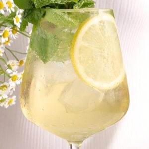 A hugo spritz mocktail with ice cubes, a slice of lemon, and a garnish of fresh mint leaves. White flowers and extra lemon slices decorate the background.