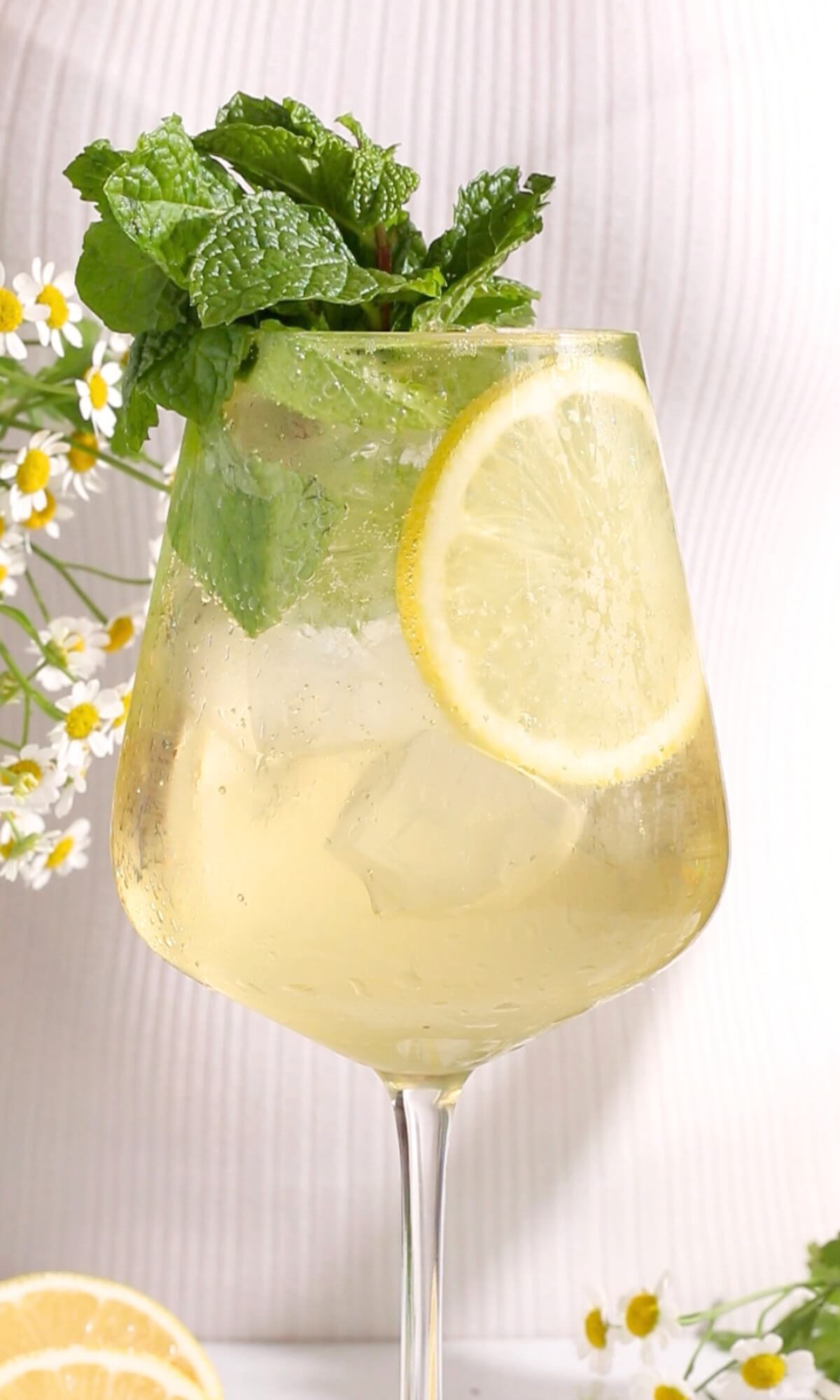 A hugo spritz mocktail with ice cubes, a slice of lemon, and a garnish of fresh mint leaves. White flowers and extra lemon slices decorate the background.