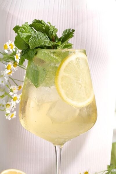 A glass of light-colored Hugo Spritz mocktail garnished with a lemon slice and fresh mint, set against a soft white background with small white and yellow flowers.