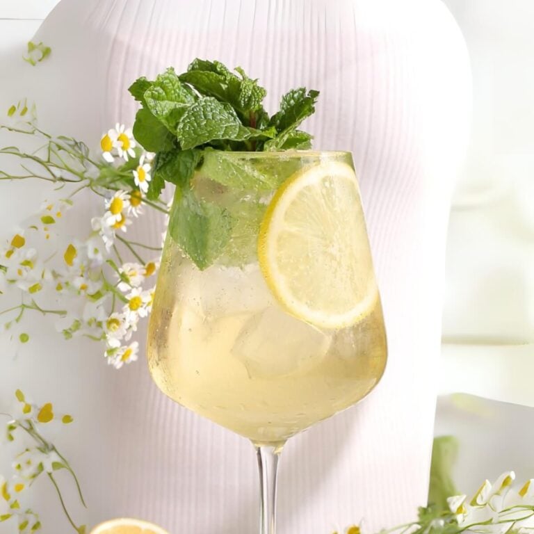 A glass of light-colored Hugo Spritz mocktail garnished with a lemon slice and fresh mint, set against a soft white background with small white and yellow flowers.