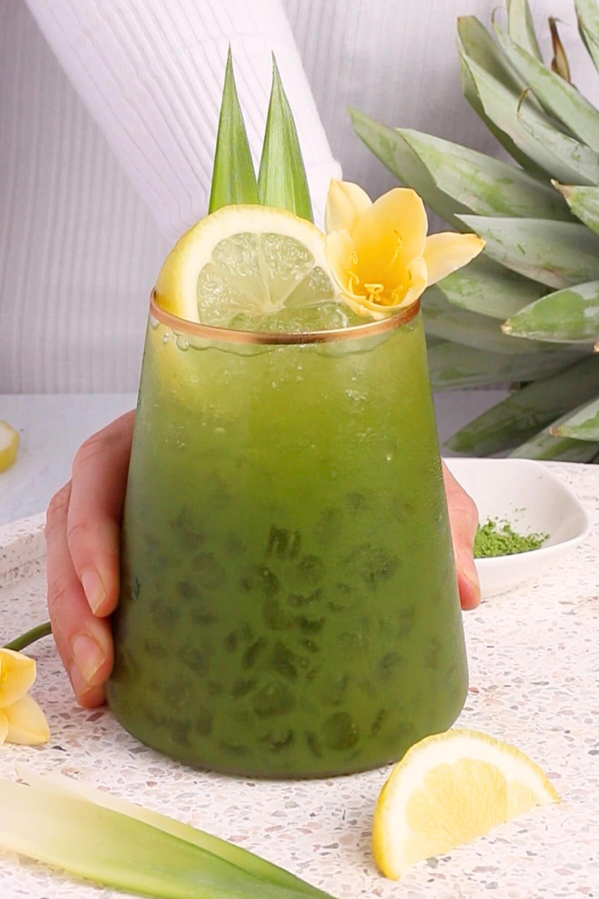 A hand holds a glass of iced green matcha drink garnished with lemon slices, a yellow flower, and pineapple leaves. Pineapple and lemon pieces are on the speckled countertop. A white plate is in the background.
