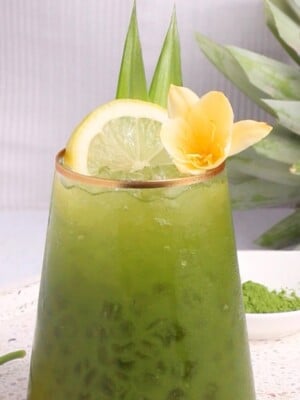 A glass of iced green matcha drink garnished with a lemon slice, two upright pineapple leaves, and a yellow flower, with a fresh pineapple and matcha powder in the background.