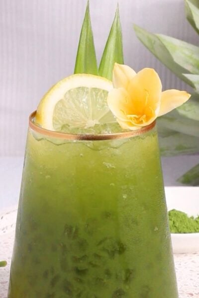 A glass of iced green matcha drink garnished with a lemon slice, two upright pineapple leaves, and a yellow flower, with a fresh pineapple and matcha powder in the background.
