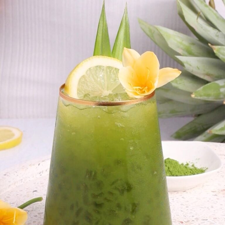 A glass of iced green matcha drink garnished with a lemon slice, two upright pineapple leaves, and a yellow flower, with a fresh pineapple and matcha powder in the background.