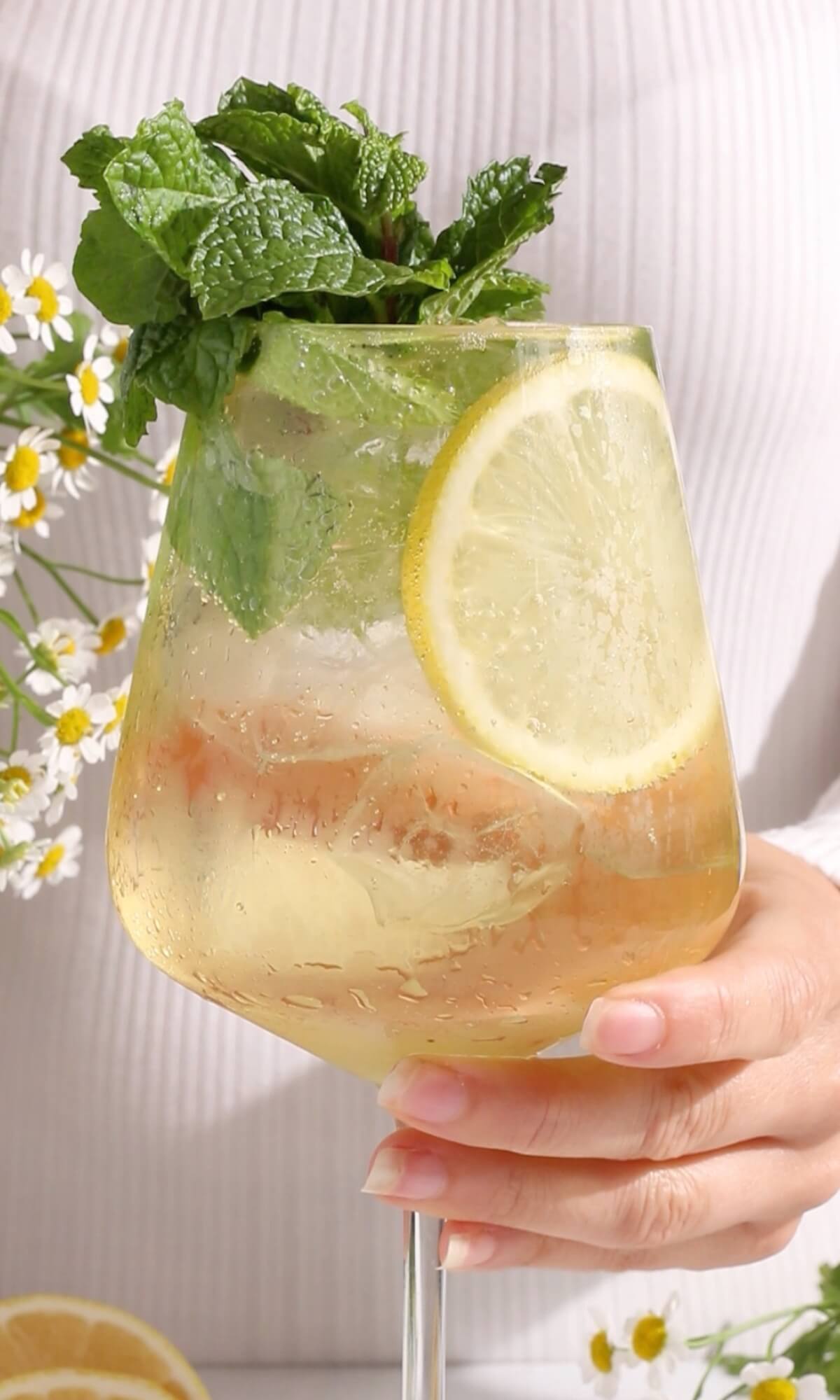 A hand holds a glass filled with a light, fizzy non-alcoholic Hugo Spritz, garnished with a lemon slice and fresh mint. Small white flowers and lemon slices decorate the background.