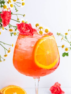 A vibrant orange cocktail in a stemmed glass, garnished with a large orange slice, red carnation, and small white flowers. More flowers are arranged around the glass against a white background.