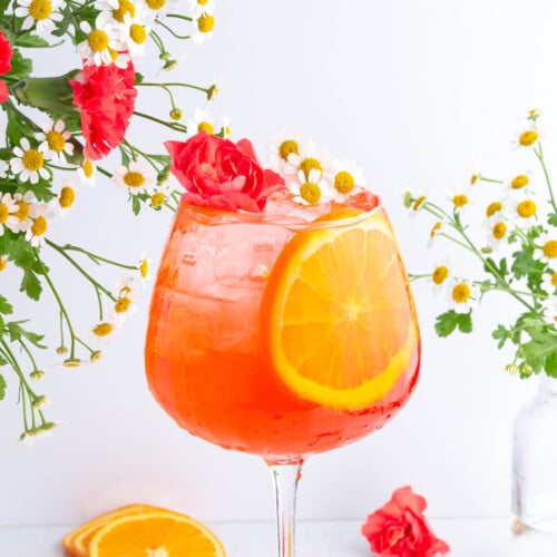 A vibrant orange cocktail in a stemmed glass, garnished with a large orange slice, red carnation, and small white flowers. More flowers are arranged around the glass against a white background.