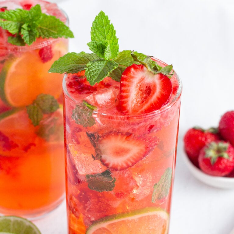 A refreshing strawberry mocktail in a tall glass, garnished with fresh strawberries, lime slices, mint leaves, and ice cubes. Another similar drink and a bowl of strawberries are in the background on a white surface.