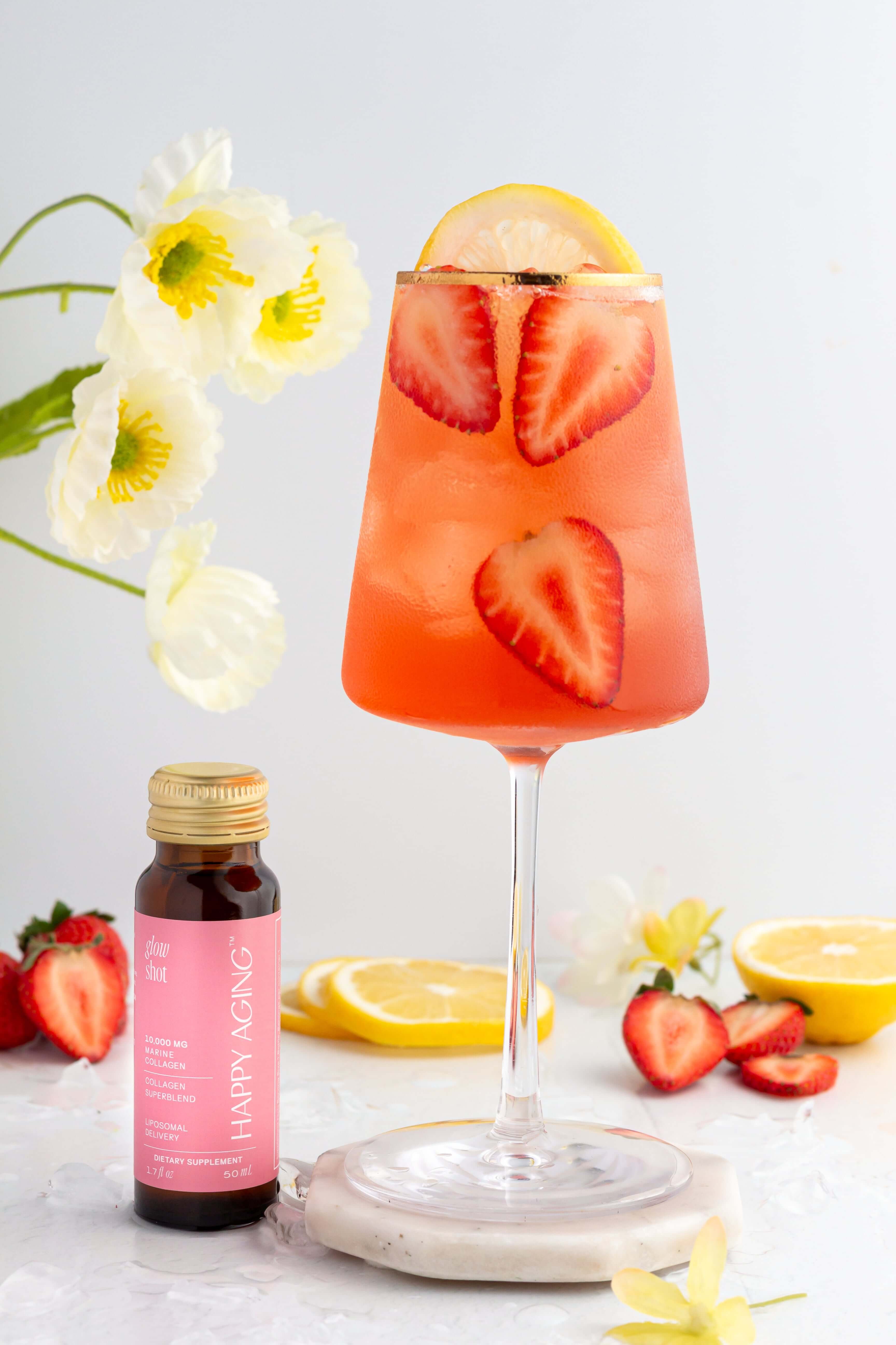 A glass of pink strawberry and lemon drink with strawberry slices and a lemon wedge, next to a small bottle labeled Happy Aging. Fresh strawberries, lemon slices, and white flowers are arranged around the scene.