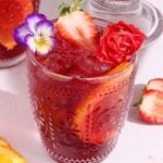 A vibrant red drink with ice, orange slices, and strawberries in an ornate glass, garnished with edible flowers and a red carnation. More fruit and flowers are scattered on the light-colored surface around the glass.