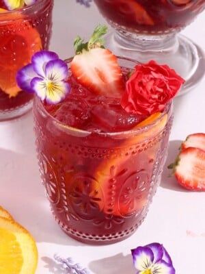 A vibrant red drink with ice, orange slices, and strawberries in an ornate glass, garnished with edible flowers and a red carnation. More fruit and flowers are scattered on the light-colored surface around the glass.